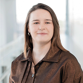 A smiling woman wearing a brown shirt stands in front of a bright university corridor.